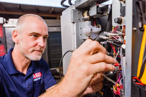Installation A concentrated male technician with a beard, wearing a dark blue ASD polo shirt, is installing electrical components within a large gray machine. He's manipulating cables and examining the connections closely. The focus is on his hands and the complexity of the task at hand.